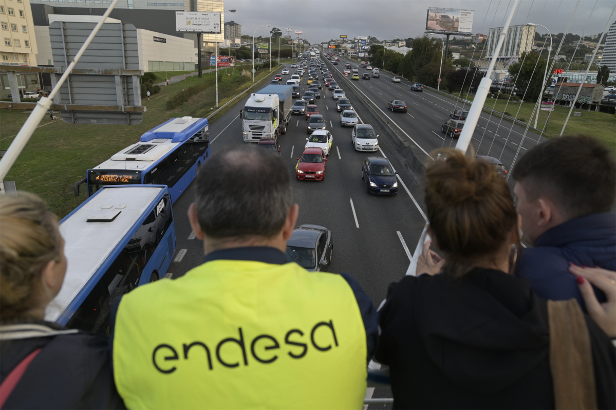 Varias persoas, unha cun chaleco de 'Endesa', observan unha caravana de automóbiles e camións mobilizada pola paralización da central térmica de Endesa en de As Pontes diríxese desde esa l